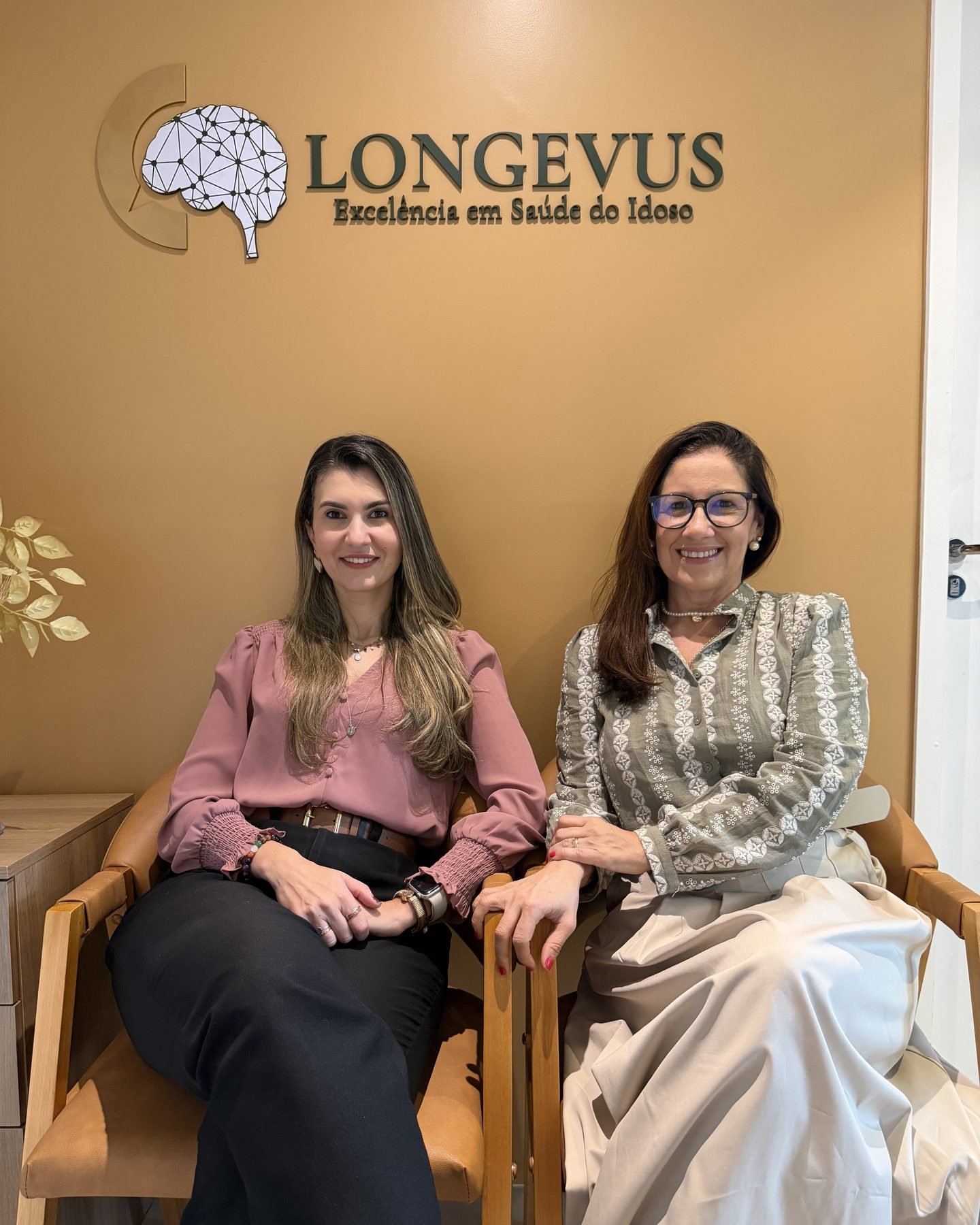 Livià Ribeiro e Débora Gomes sentadas na recepção da Longevus, em frente à parede com o logotipo da clínica.